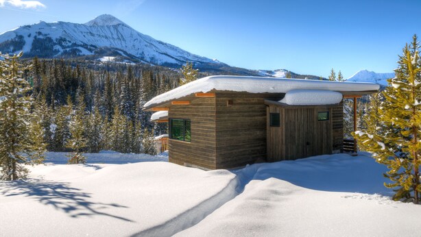 Une cabane en bois, dont le toit est recouvert de neige, entourée d’un paysage enneigé et d’arbres, avec une montagne en arrière-plan.