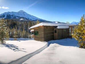 Une cabane en bois, dont le toit est recouvert de neige, entourée d’un paysage enneigé et d’arbres, avec une montagne en arrière-plan.
