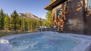 Un bain à remous avec une terrasse en bois et des chaises, entouré d’arbres et avec une montagne en arrière-plan.