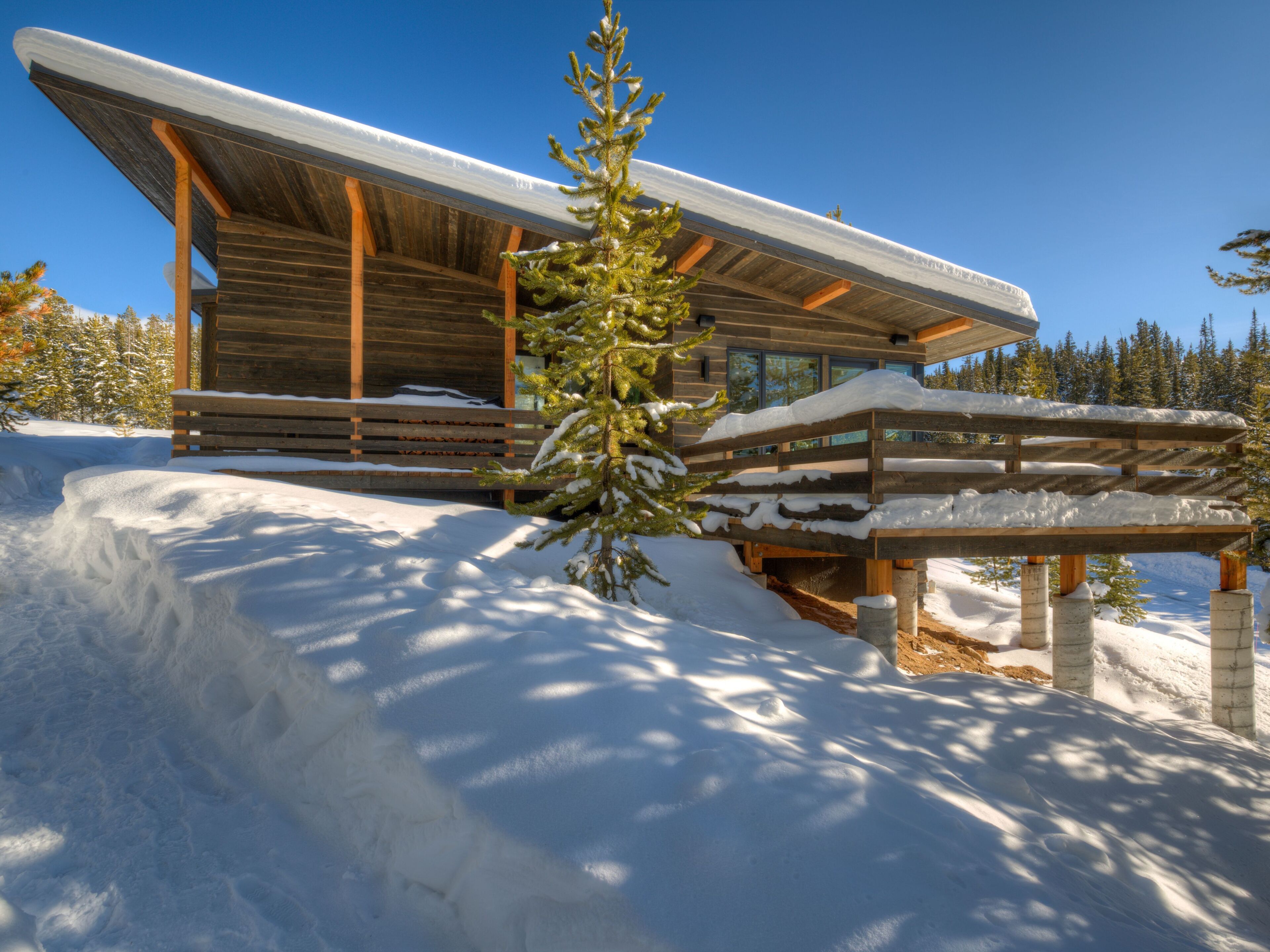 Une cabane en rondins moderne, dotée d’une grande terrasse et d’un porche abrité, entourée de neige et d’arbres.