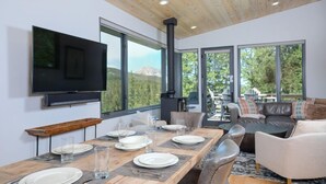 Un espace repas moderne avec une table en bois dressée pour quatre personnes, un grand téléviseur à écran plat fixé au mur et une grande fenêtre offrant une vue sur un paysage montagneux.