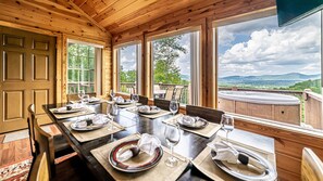 Un coin repas avec une table dressée pour six personnes, de grandes fenêtres offrant une vue panoramique sur les montagnes et un jacuzzi visible à travers les fenêtres.