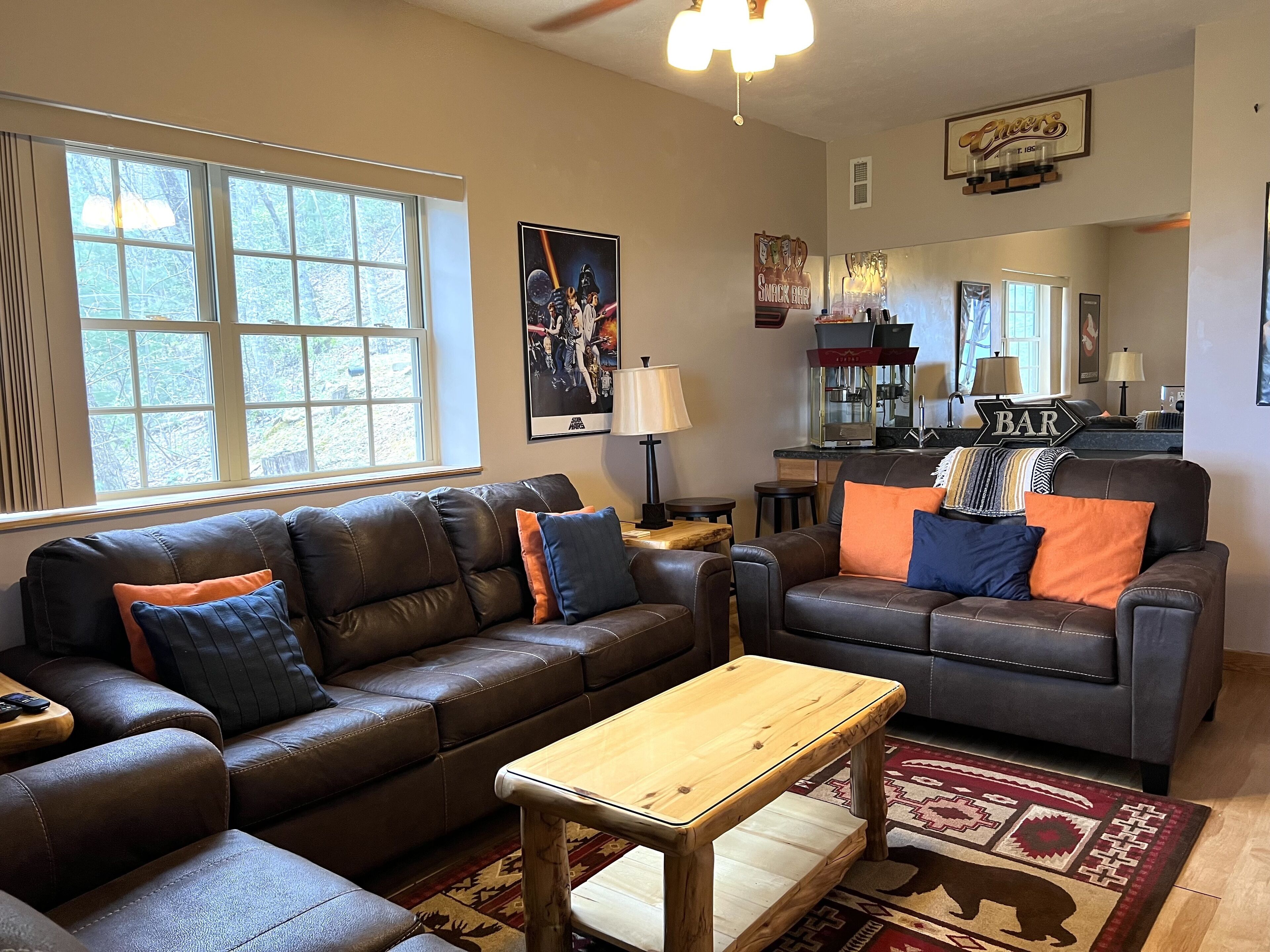 A living room with leather sofas, a wooden coffee table, and a rug. There is a bar area with a 'BAR' sign and a 'SNACK BAR' sign.