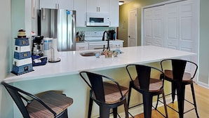 A modern kitchen with white cabinets, a stainless steel refrigerator, a microwave, and a sink. There are three black bar stools with wooden seats. - Sea Colony 5A | Oceanfront Pool + Beach Walkway! (Carolina Beach)