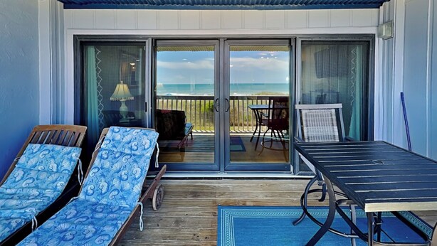 A covered outdoor area with two lounge chairs, a table, and a view of the beach through glass doors. - Sea Colony 5A | Oceanfront Pool + Beach Walkway! (Carolina Beach)
