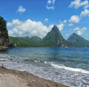 A beach with clear water, rocky shore, and green mountains in the background.
