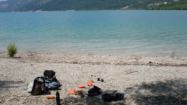 A pebbled beach with bags and gear, a clear lake, and a forested hill in the background. - Close to the Gorges du Verdon Apartment with garden, terrace, panoramic view (Puimoisson)