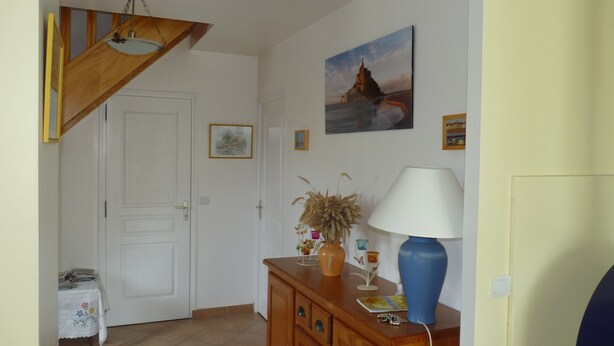 Een hal met een houten trap, een witte deur, een klein tafeltje met een lamp en een wand met ingelijste foto's.