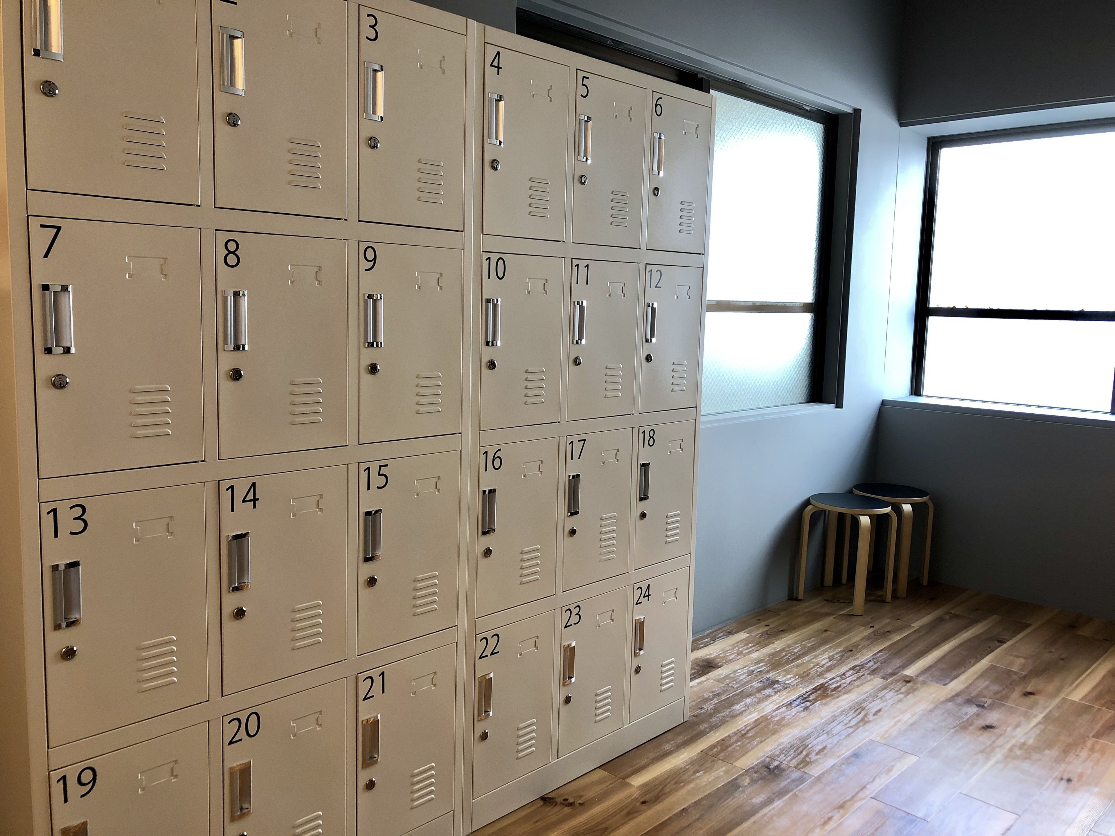 A row of beige lockers numbered 1-24, wooden flooring, and a window with blinds.