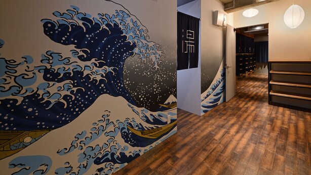 A hallway with a large wave mural on the wall, wooden flooring, and a door leading to another room. - Santiago Guesthouse Naha - Hostel (Naha)