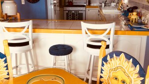Une cuisine avec des meubles blancs, une cuisinière et un évier. On y trouve deux chaises blanches et un tabouret bleu.