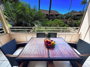 A wooden table with a fruit bowl, surrounded by wicker chairs, on a balcony with a view of a building and greenery.