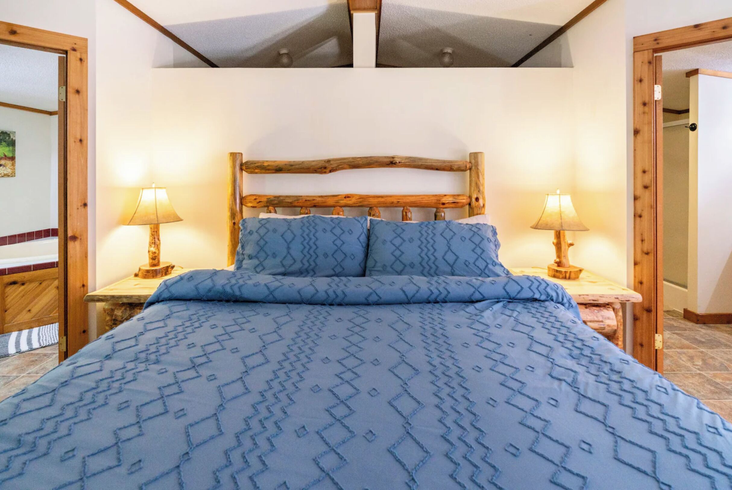 A bedroom with a wooden headboard, a patterned blue bedspread, two bedside lamps, and a wooden nightstand.