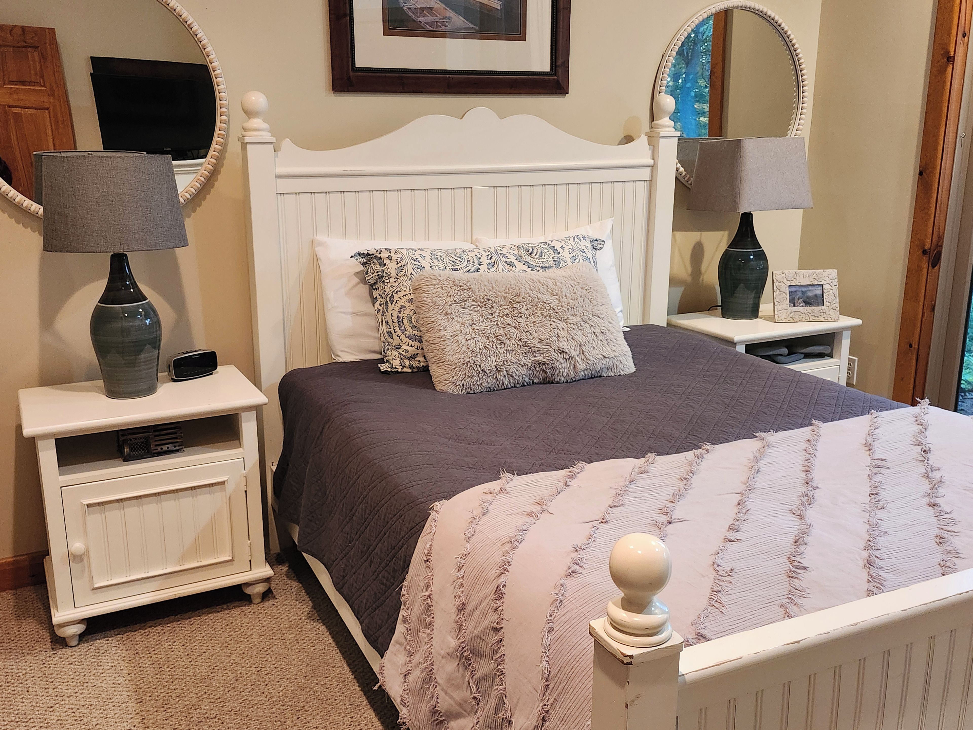 A bedroom with a white bed, two bedside tables, a lamp, a television, and a framed picture on the wall.