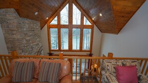 Interior acogedor de una cabaña con pared de piedra, techo de madera y ventanas amplias que ofrecen vistas al exterior.