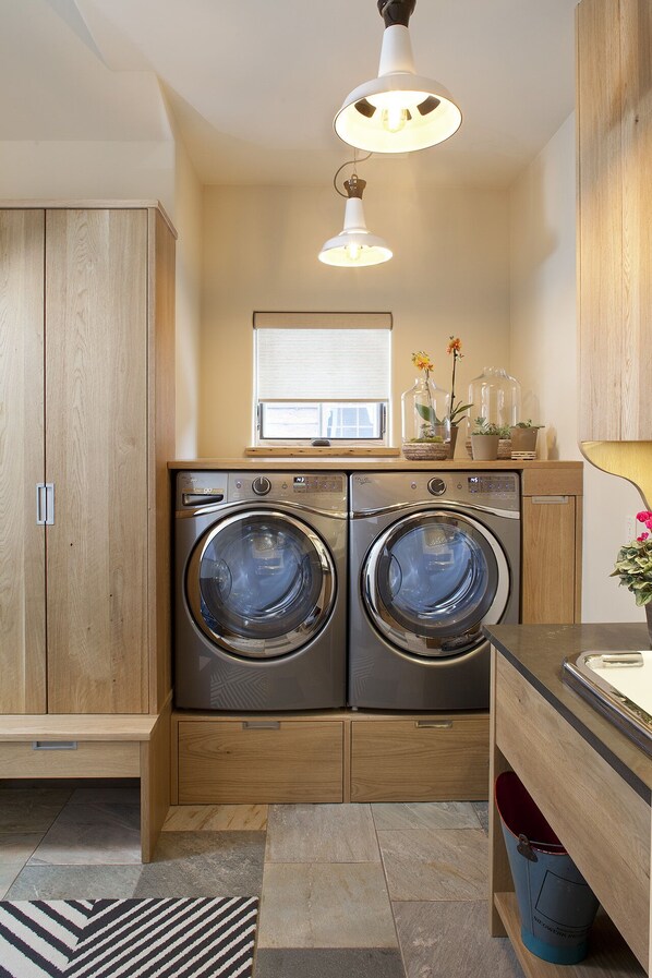 A laundry room with two front-loading washing machines, wooden cabinets, a window with blinds, and a hanging light fixture. - Come Home To Tomorrow's Comforts in Historic CB! Book for summer soon!! (Crested Butte)
