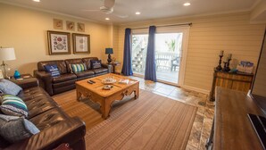 A living room with a brown leather sofa, a wooden coffee table, and a dining table with a lamp. There are framed pictures on the wall and a sliding glass door leading to an outdoor area. -  Island's End Welcomes You to Paradise!   (Pensacola Beach)