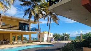 A pool area with a curved edge, palm trees, and a building with a balcony. - Caribbean Beach Front Gem 2 (Punta Pozuelo, Guyama)