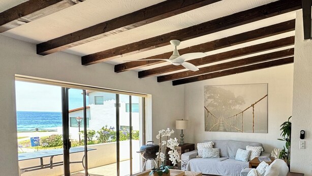 A spacious living room with a large sliding glass door leading to a balcony with a view of the ocean. There is a gray sofa, two gray ottomans, and a wooden coffee table.