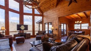 A cozy cabin interior with a wooden interior, leather furniture, and large windows offering a view of the outdoors. - 5 Suite Toccoa River Lodge Sleeps 12 | Hot Tub + Game Room | Fishing Access (Blue Ridge)