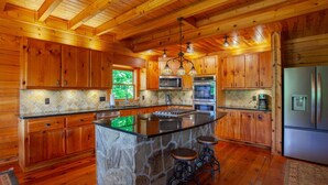 A spacious kitchen with wooden cabinets, a central island with stools, a stone accent wall, and modern appliances. - 5 Suite Toccoa River Lodge Sleeps 12 | Hot Tub + Game Room | Fishing Access (Blue Ridge)