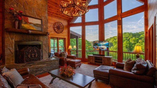 A spacious living room with a stone fireplace, leather sofas, a large TV, and a scenic view through large windows. - 5 Suite Toccoa River Lodge Sleeps 12 | Hot Tub + Game Room | Fishing Access (Blue Ridge)