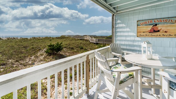 Un balcone sul mare con vista sulla spiaggia e l'oceano, dotato di un pavimento in legno, sedie bianche e un tavolo con una lampada.