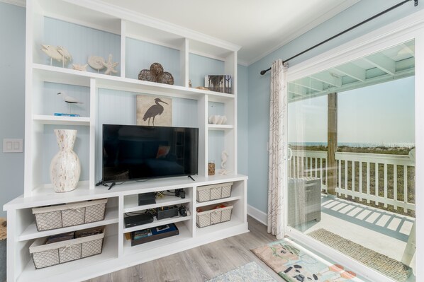 A living room with a TV on a white entertainment unit, storage bins, and a view of the beach through a glass door.