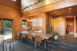 A wooden interior with a dining area featuring a long table and chairs, a chandelier, and a potted plant.