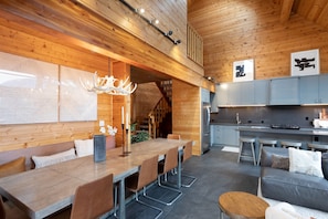 A modern interior with a wooden ceiling, a dining area with a long table and chairs, a kitchen with grey cabinets, and a staircase leading to an upper level.