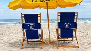 Two beach chairs under a yellow umbrella with 'Pleasure Island Rentals' branding and contact number.