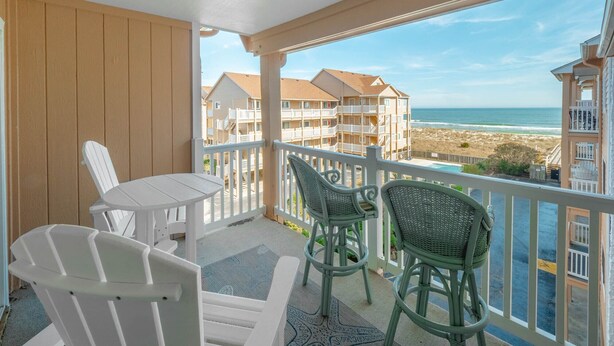 A balcony with a view of the beach and ocean, white chairs, and a small table.