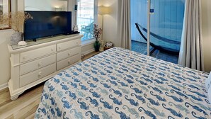 A bedroom with a bed, a TV, a dresser, a window with blinds, a chair, and a balcony with a view.