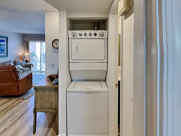 A compact laundry room with a washer and dryer, wooden flooring, a small table, and a living area with a sofa and a clock. - Sand Pebbles C21 | Ocean View + Community Pool! (Carolina Beach)