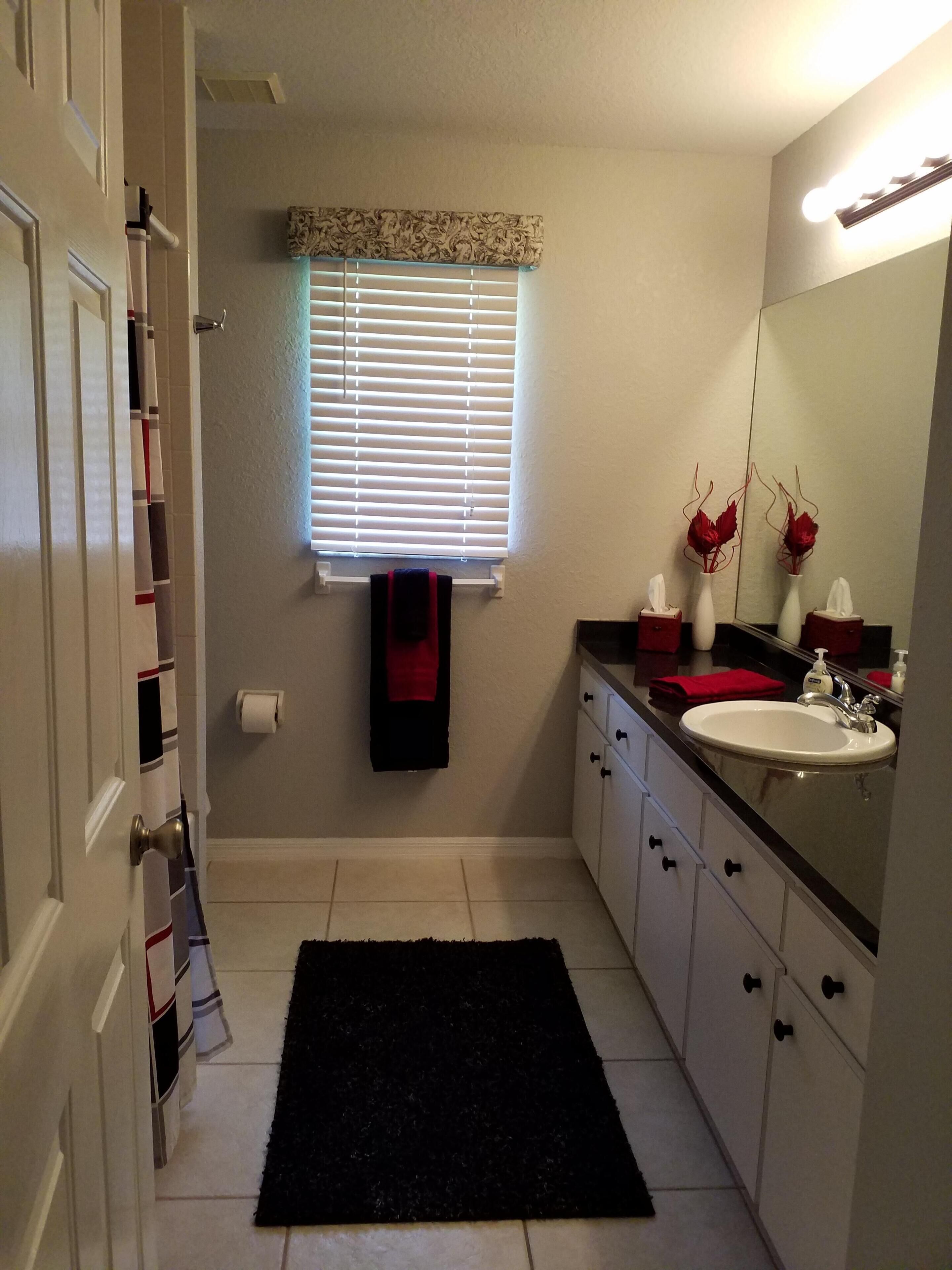 Une salle de bain avec un lavabo blanc, un miroir, un tapis noir, un porte-serviettes et une fenêtre avec des stores.