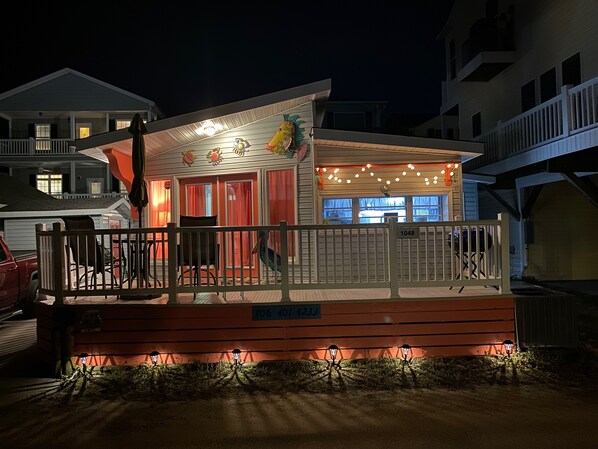 A small, brightly lit building with a porch, a welcome sign, and a number sign.