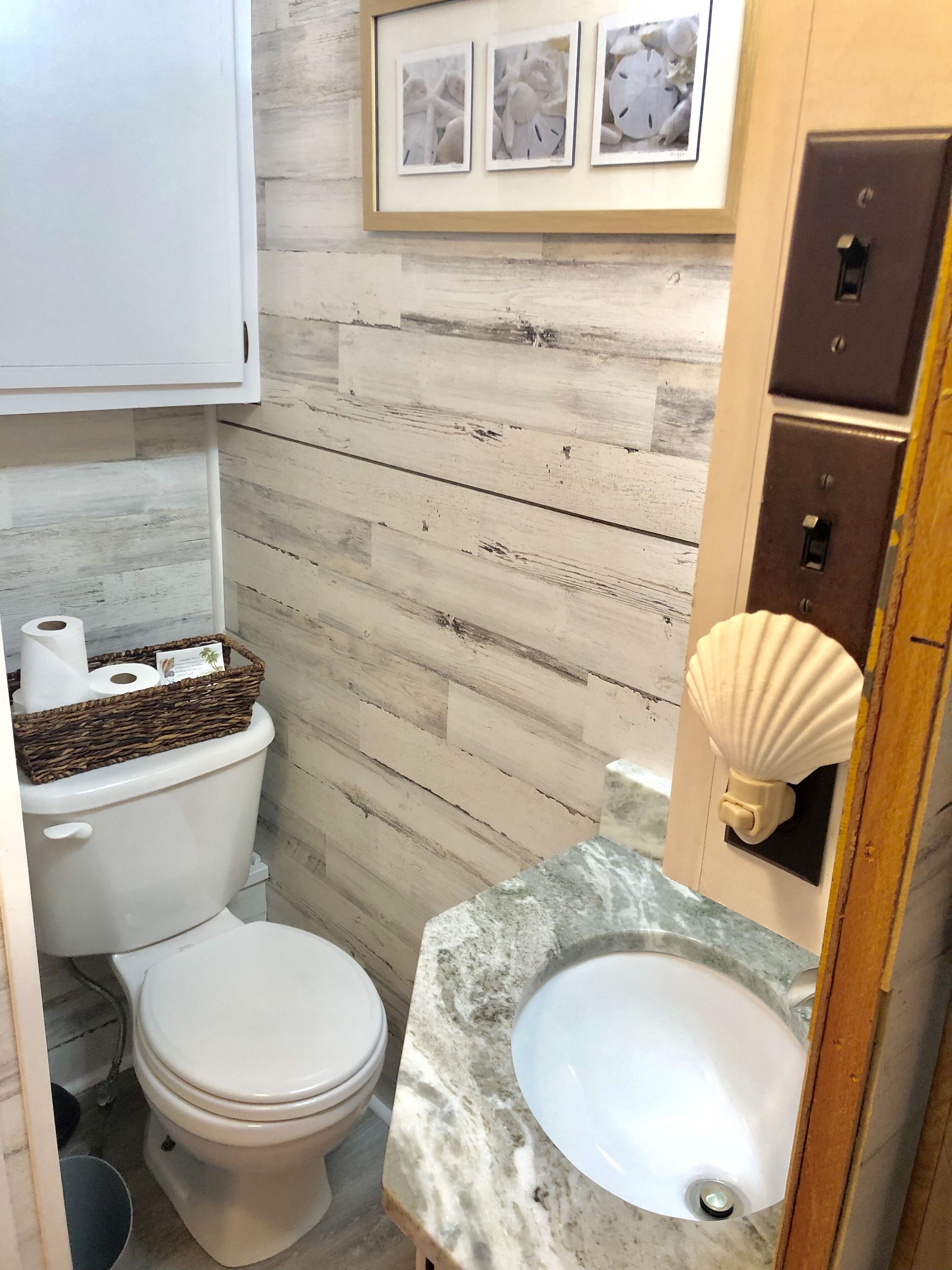 A bathroom with a toilet, sink, and wooden wall paneling. There are framed pictures on the wall and a basket of toiletries on the toilet.