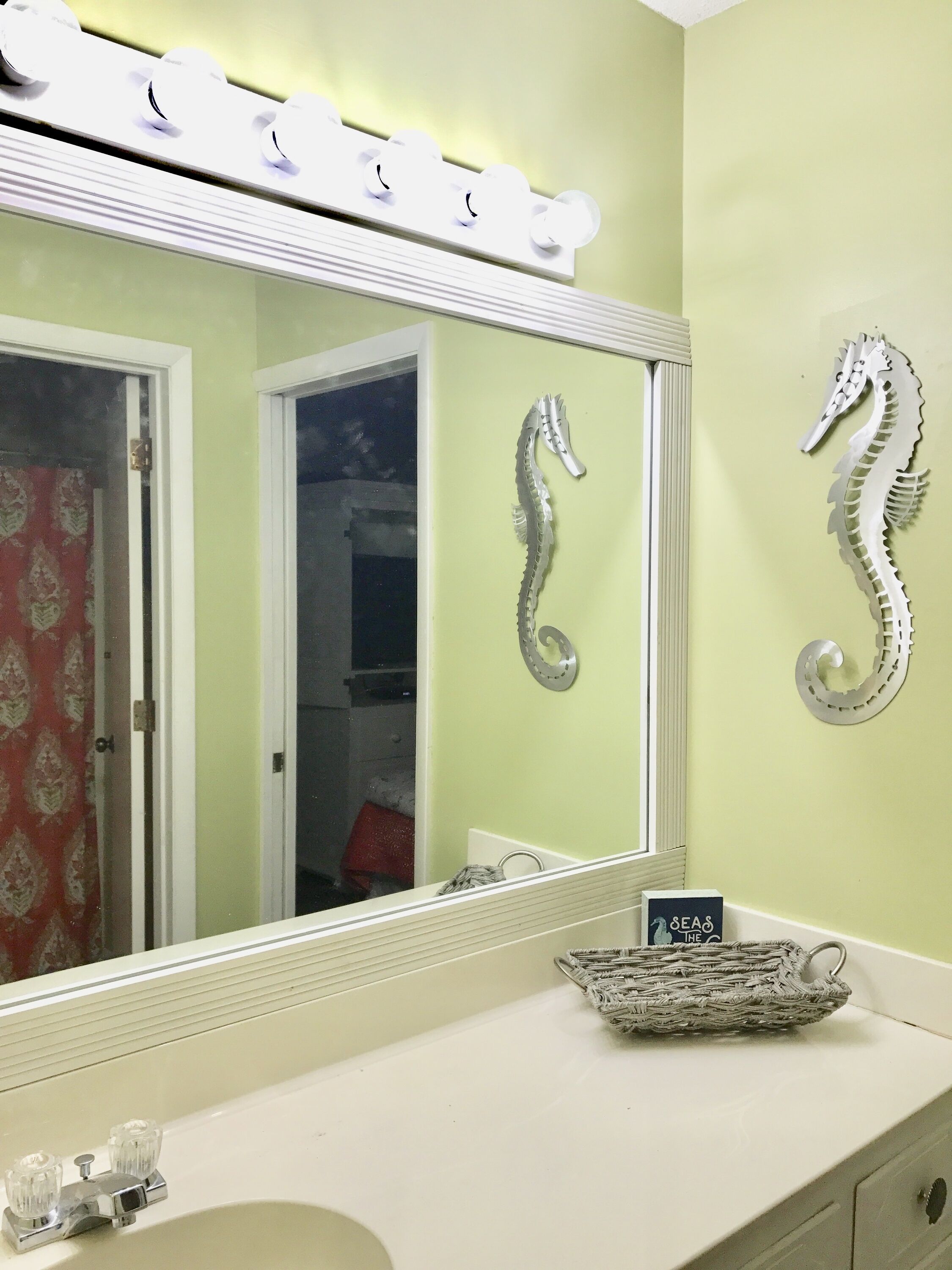 A bathroom with a vanity, a mirror with lights, a seahorse decoration, and a sign that says "SEAS THE".