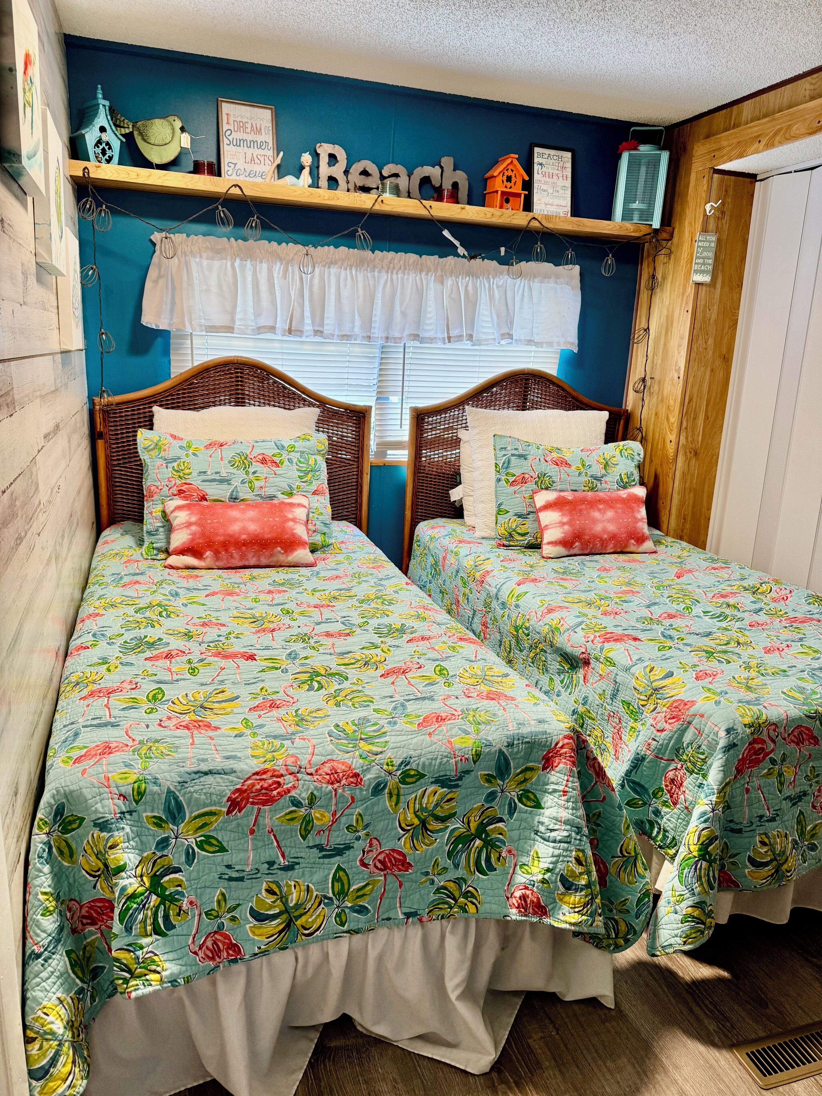 Une chambre avec deux lits, chacun agrémenté de draps et d’oreillers à motifs floraux assortis. Une étagère au-dessus des lits est décorée de divers objets et porte une inscription indiquant « Plage ».
