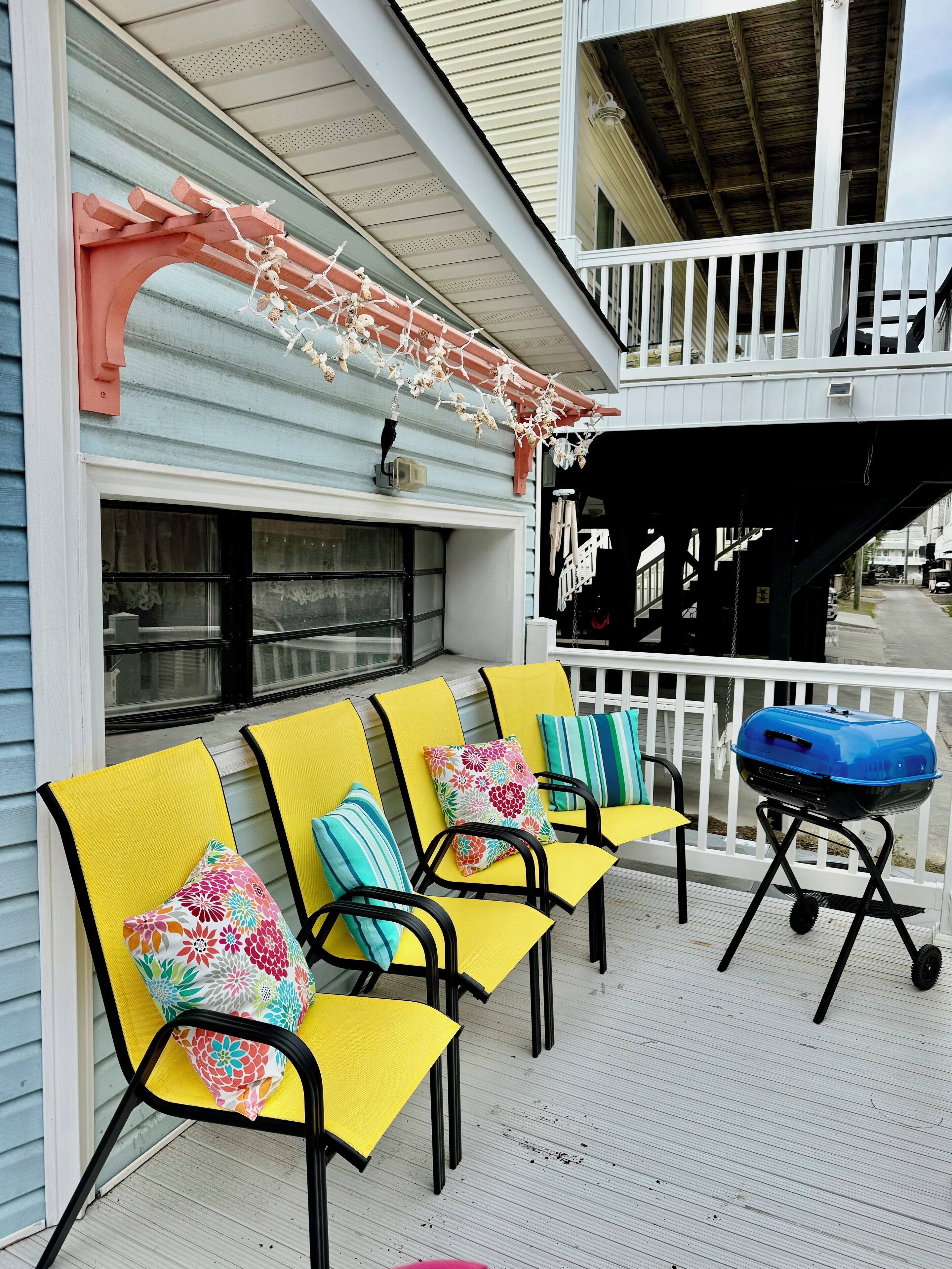 A covered outdoor seating area with yellow chairs and colorful cushions, a barbecue grill, and a white railing.