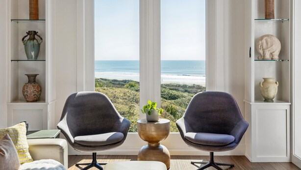 Una habitación con vistas a la playa, dos sillas, una mesa con una planta y estantes con objetos decorativos.