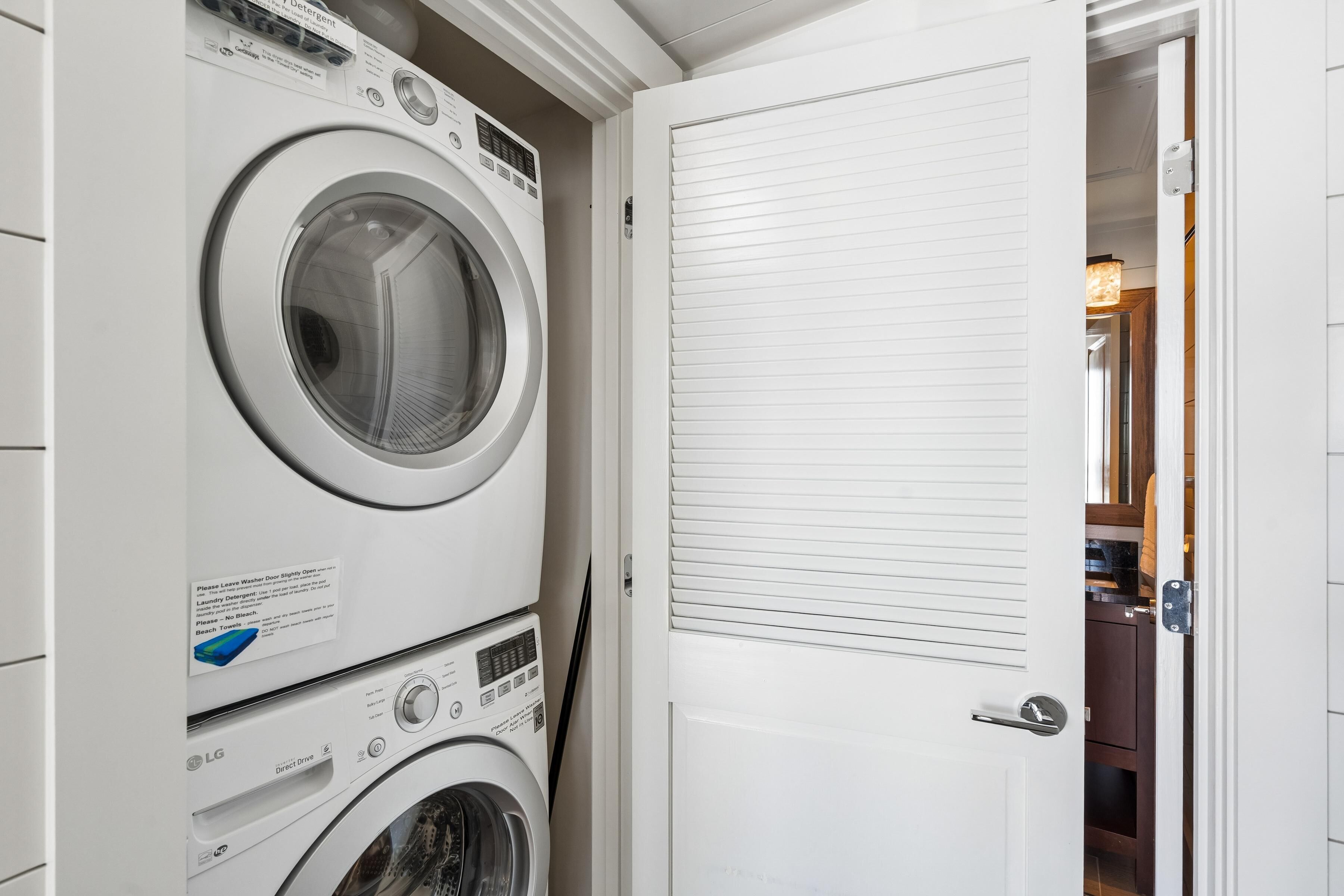 Lavandería con lavadoras y secadoras apiladas, una puerta cerrada y un pasillo visible que conduce a una habitación con piso de madera.