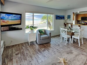 A modern living room with a large window, a flat-screen TV, a gray sofa, a dining table with white chairs, and a kitchen area.