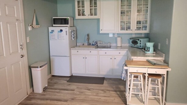 Una cocina compacta con armarios blancos, microondas, refrigerador, lavamanos, cafetera y una pequeña mesa de comedor con taburetes.
