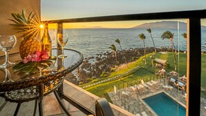 A balcony with a glass table, a pineapple, and a bottle of champagne. The view includes a pool, a lawn, and a body of water with palm trees. - 605 - Top Floor, Luxury Oceanfront, Newly-renovated with A/C!  1/1.5 (Kihei)
