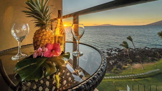 A table with a pineapple, a bottle, and two glasses, overlooking a coastal view with a sunset. - 605 - Top Floor, Luxury Oceanfront, Newly-renovated with A/C!  1/1.5 (Kihei)