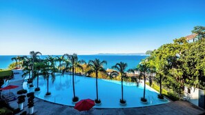A clear blue sky, a swimming pool surrounded by palm trees, and a view of the sea.