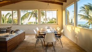 Un espace repas doté d’un plafond en bois, de grandes fenêtres donnant sur des palmiers, une table à manger avec des chaises, et un canapé avec des coussins.