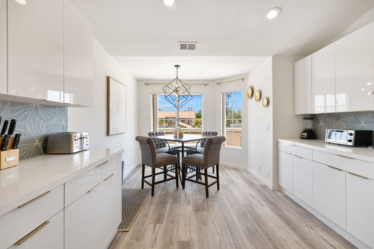 A modern kitchen with white cabinetry, a dining table with four chairs, a chandelier, and a view of the outside.
