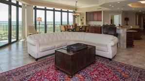 A spacious living room with a white sectional sofa, a dark wooden coffee table, and a patterned rug. There is a large window offering a view of the outdoors. - Best Prices for 4 BR Corner Unit! (Orange Beach)